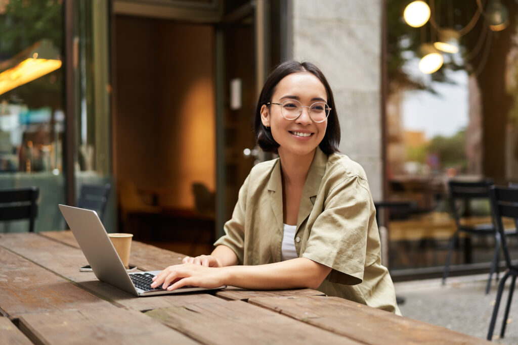 portrait-of-asian-girl-works-in-cafe-uses-laptop-and-sits-outdoors-on-street-digital-nomad-and-online-learning-concept